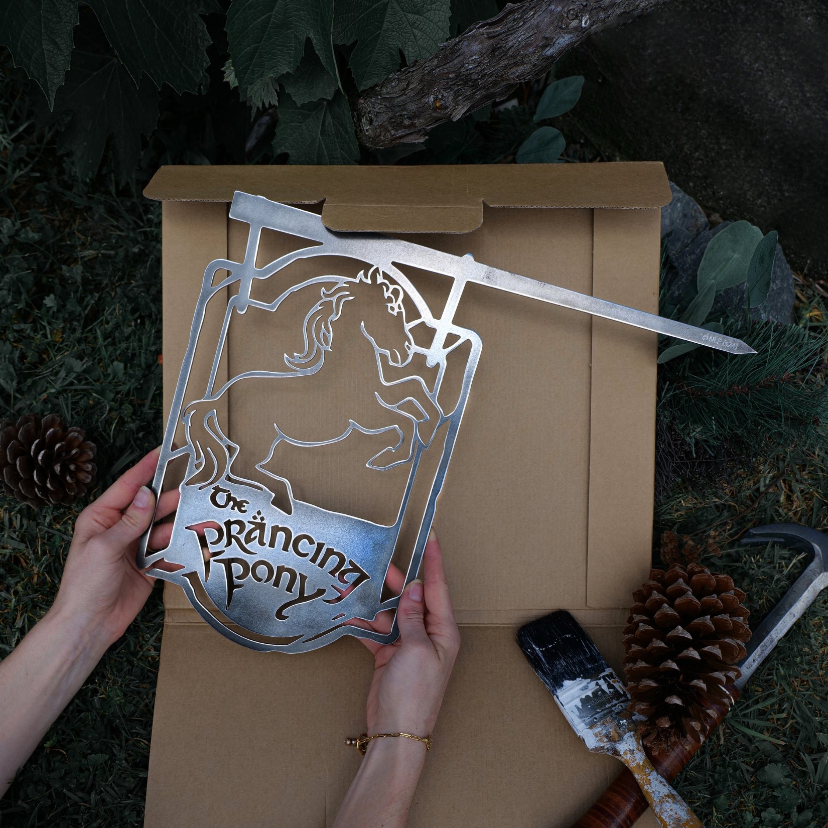 Hands hold Metalbird Canada’s THE PRANCING PONY™ metal silhouette sign over an open box outdoors, surrounded by pinecones, leaves, a paintbrush, and a hammer—ideal for adding unique yard art to your space.