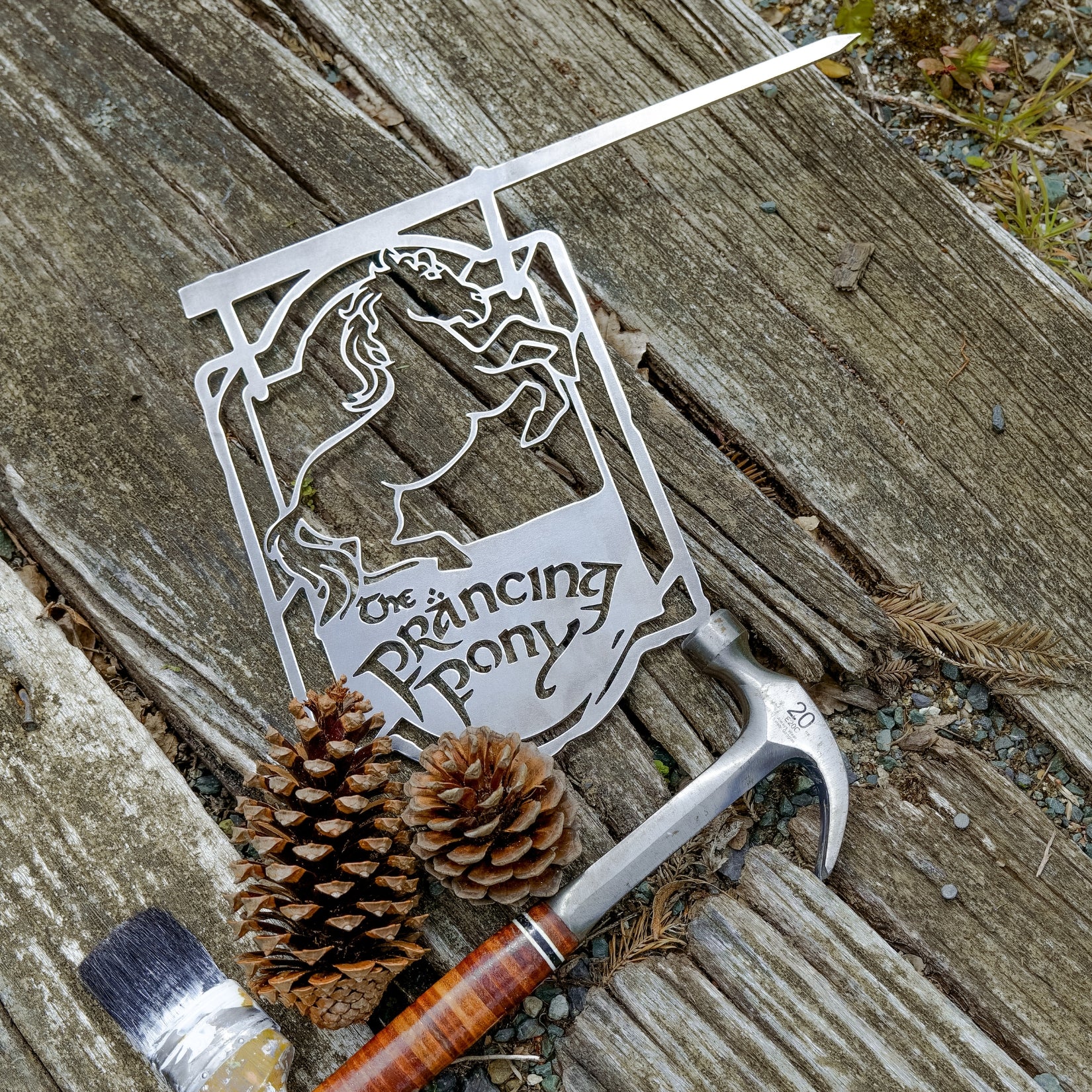A metal silhouette sign by Metalbird Canada displays a prancing horse and THE PRANCING PONY™ text on weathered wood—surrounded by a hammer, pinecones, and paintbrush—making it perfect unique garden or yard art for any outdoor space.