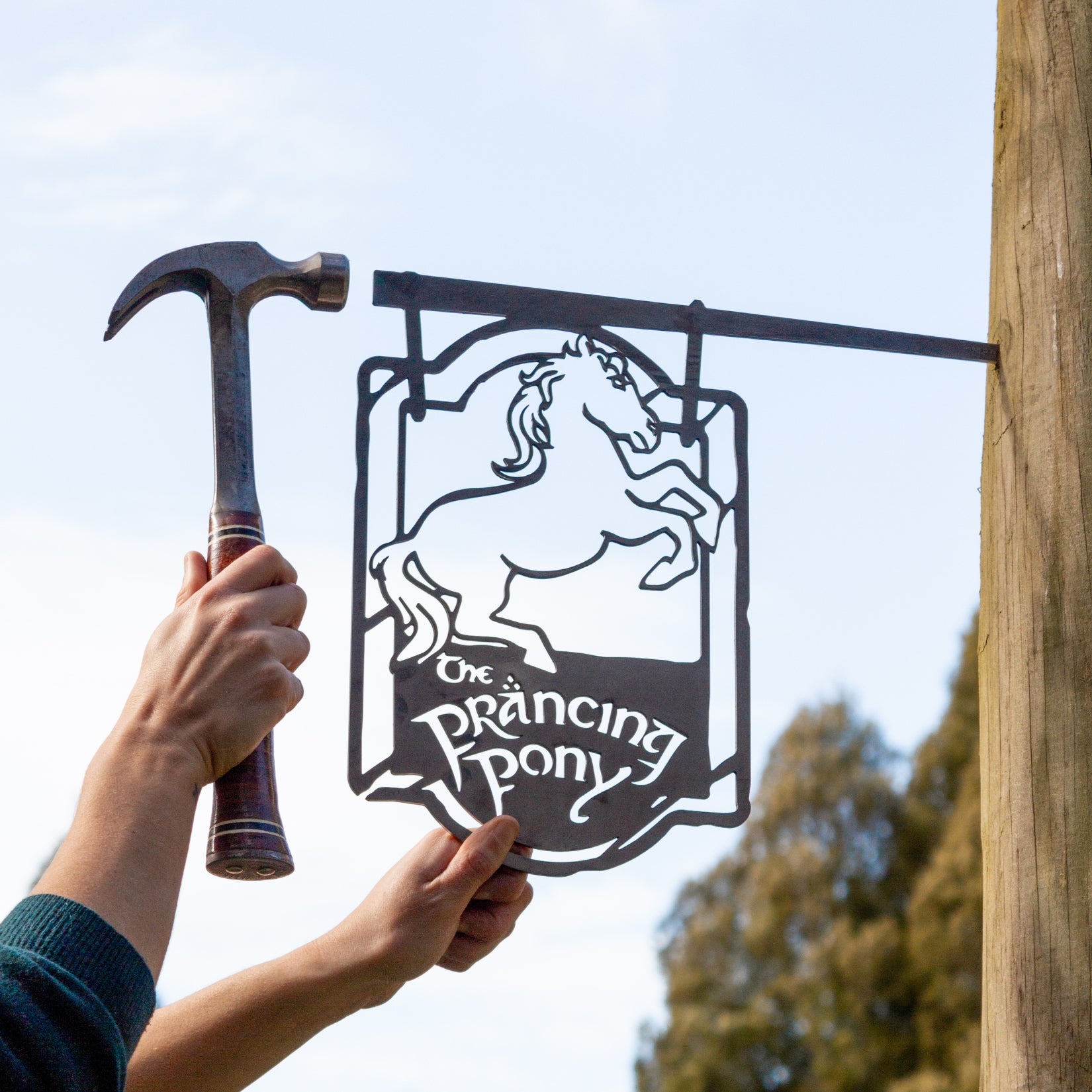 Someone holds up Metalbird Canada’s THE PRANCING PONY™ black metal sign with a rearing horse and text, while another hand hammers near a post outdoors—unique garden art and a thoughtful gift for equestrian fans.