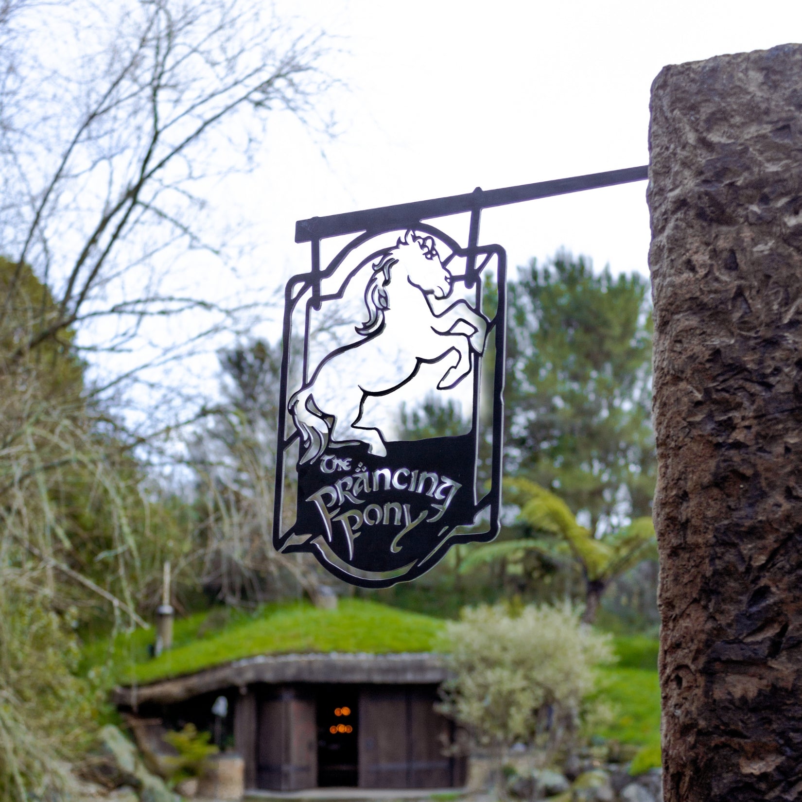 A corten steel sign from Metalbird Canada, featuring THE PRANCING PONY™, hangs outside a rustic building with greenery—an ideal gift for classic inn fans.