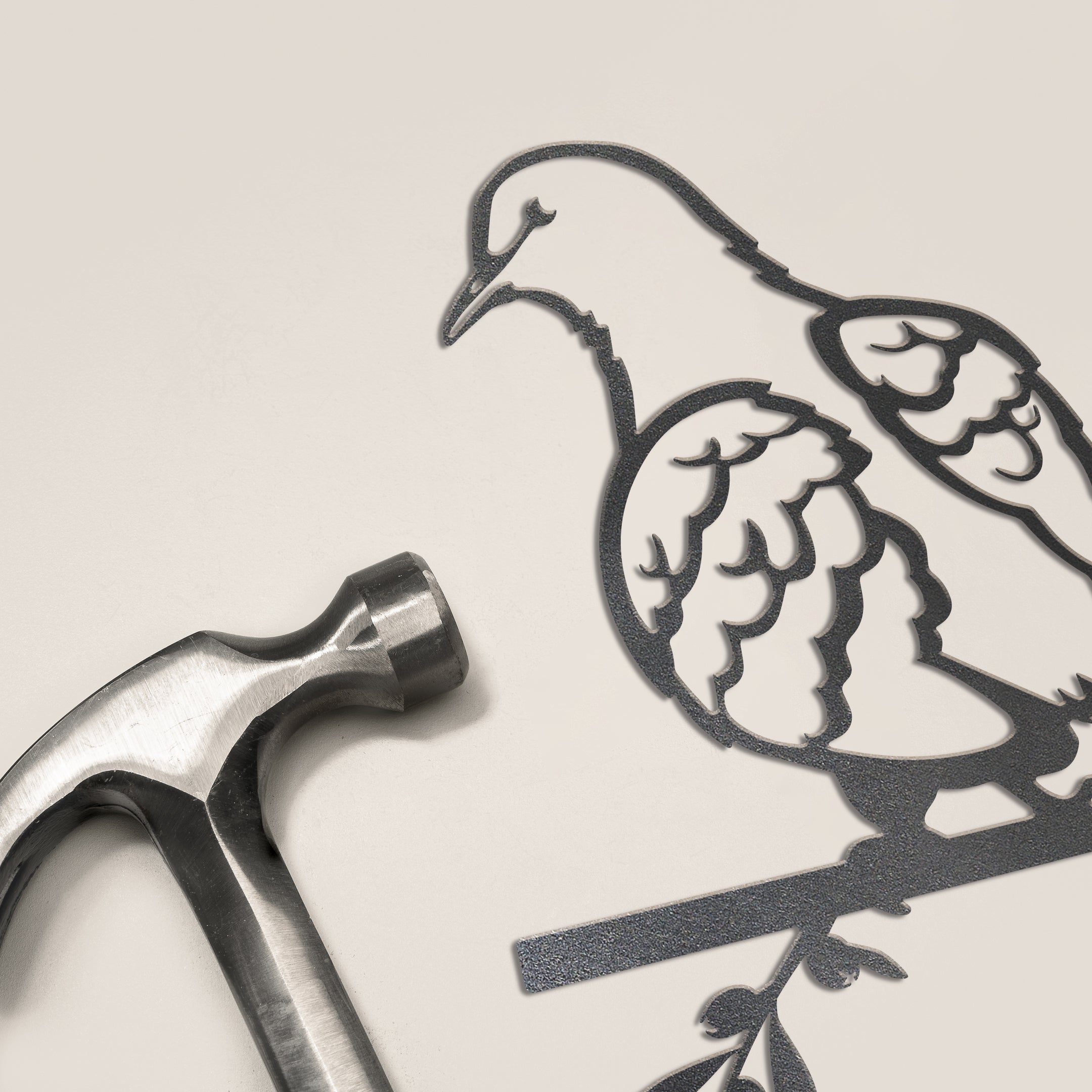A silver hammer rests beside the Metalbird Canada Turtle Dove—a minimalist, metallic outline of a bird on a branch against a light background, making for elegant yard art and a unique gift idea.