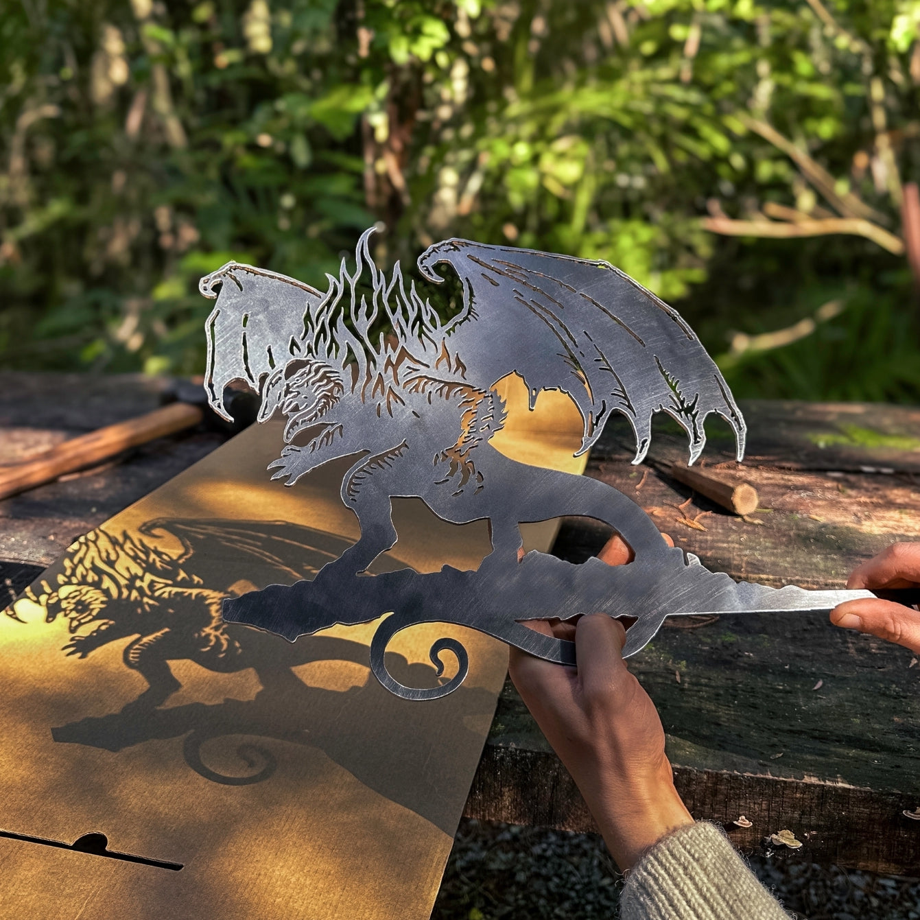 A person holds the BALROG™ metal dragon cutout by Metalbird Canada, its wings casting a striking shadow on cardboard outdoors amid lush greenery—an eye-catching piece of garden art.