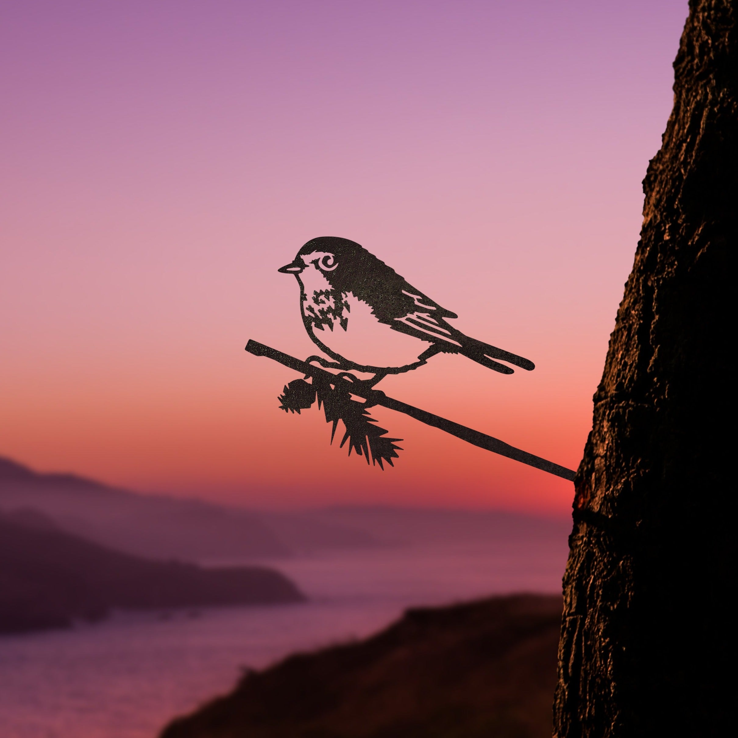 The Magnolia Warbler by Metalbird Canada is a corten steel bird silhouette on a branch, mounted on a tree trunk and set against a scenic sunset—an eye-catching piece of yard art.