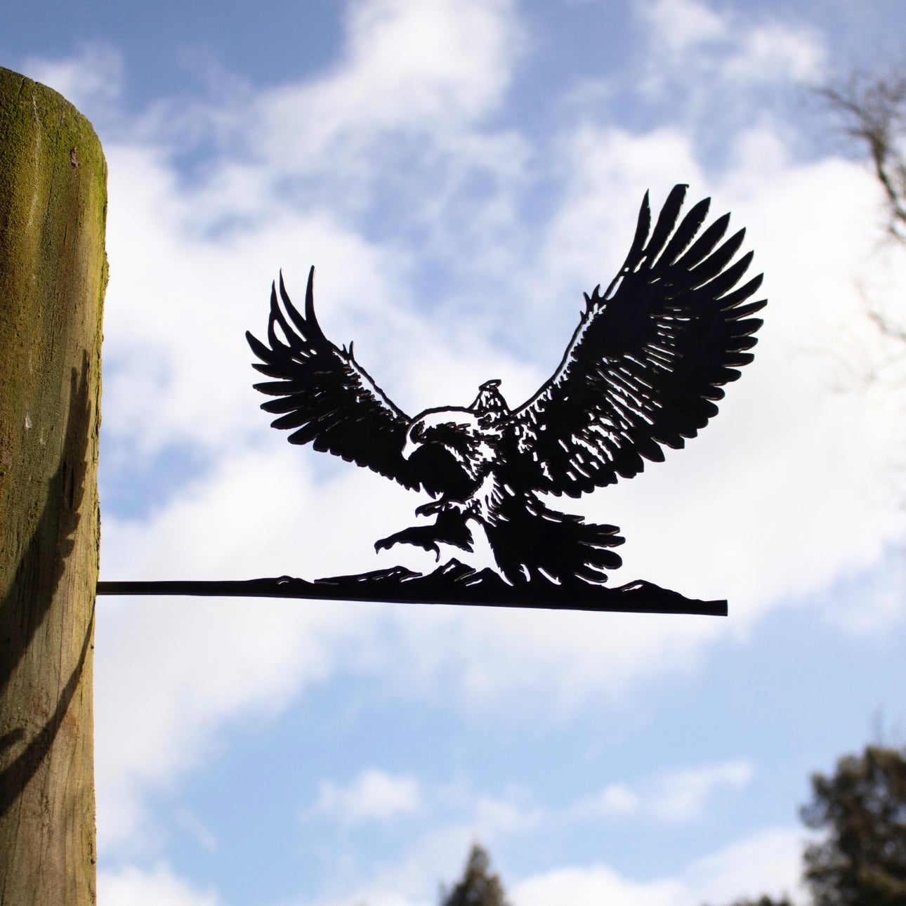 A striking black metal silhouette of GANDALF™ ON GWAIHIR™ by Metalbird Canada, mounted on a wooden post against a blue sky with white clouds—unique yard art with trees adding depth in the background.