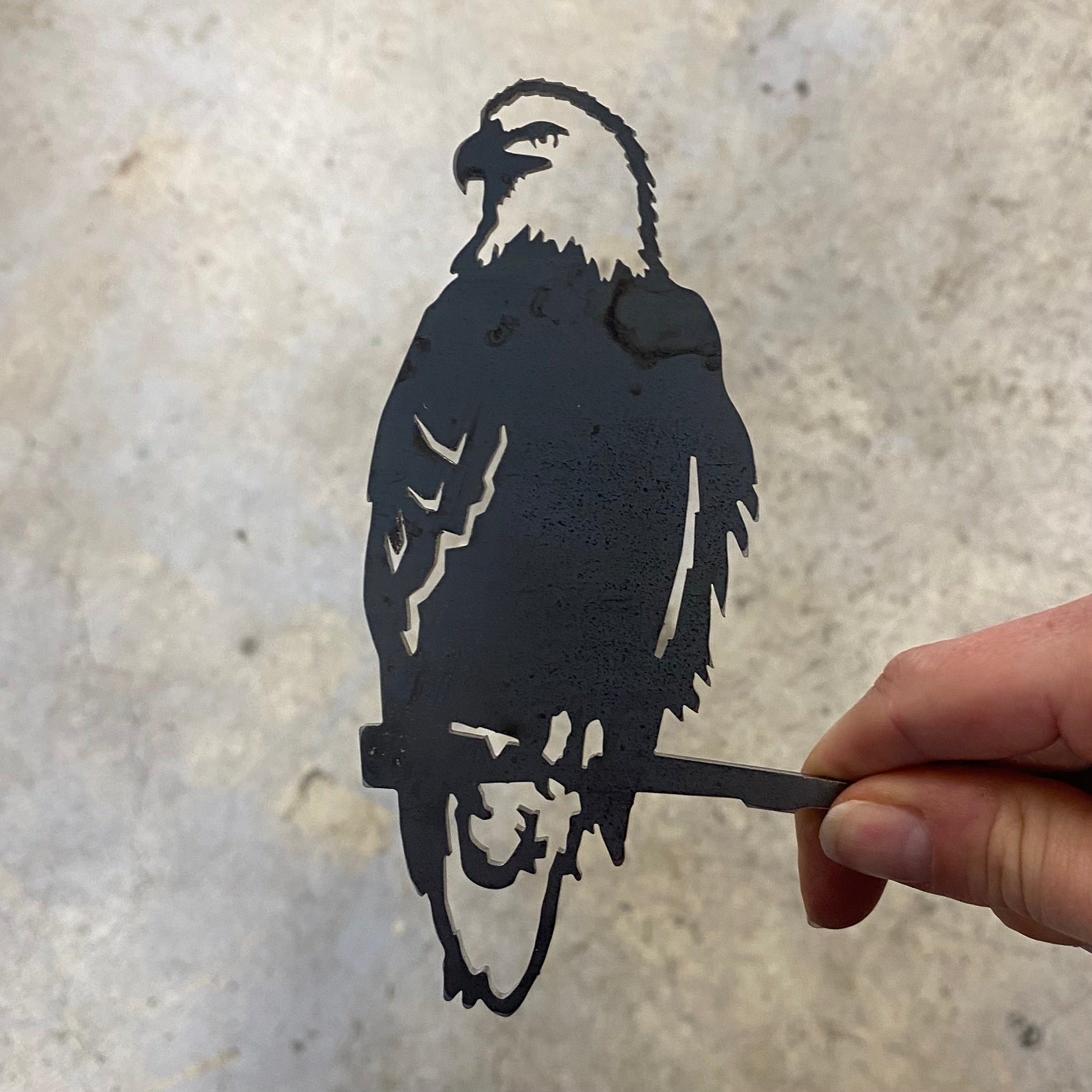 A hand holds the Mini Eagle by Metalbird Canada, a corten steel cutout showing an eagle perched on a branch with detailed feathers and beak. The bold silhouette contrasts sharply against a plain, light background.