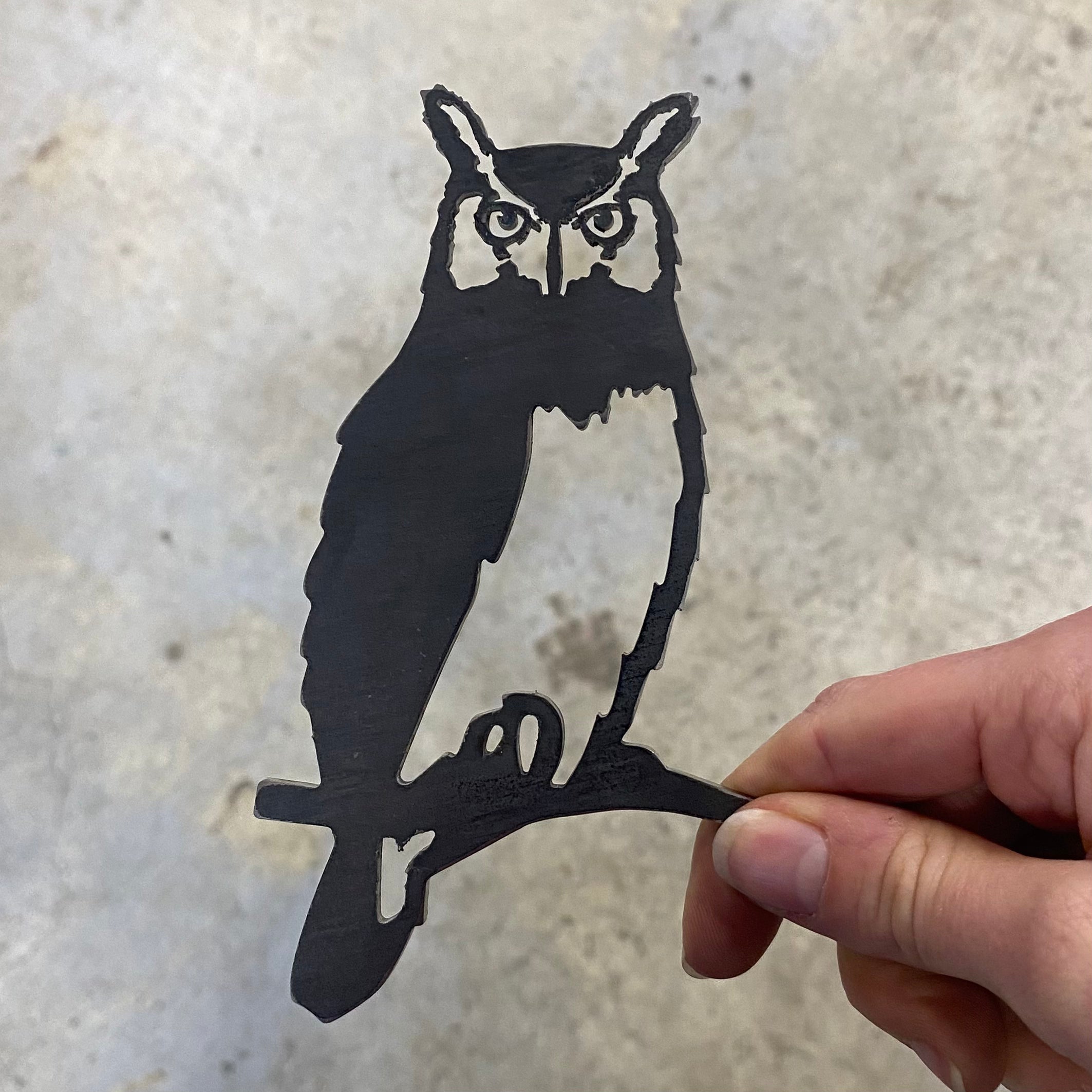 A hand holds Metalbird Canada's Mini Owl, a flat black metal silhouette perched on a branch—ideal as garden art—set against a light gray, textured background.