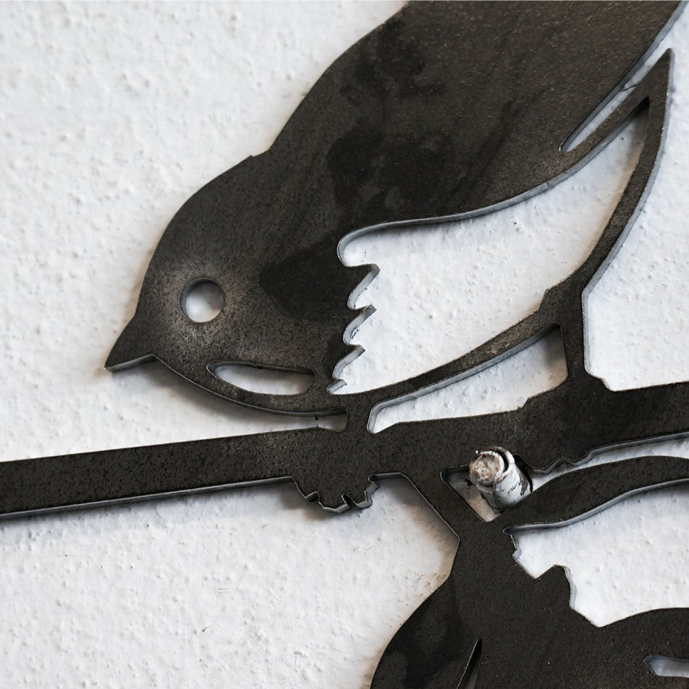 A close-up of Metalbird Seconds by Metalbird Canada - Seconds, a black corten steel wall art shaped like a bird on a branch, mounted on a textured white wall—an ideal unique gift.