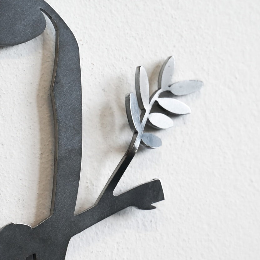 A close-up of the Metalbird Seconds by Metalbird Canada - Seconds shows a corten steel wall art piece featuring part of a branch with flat, oval leaves, creating an elegant silhouette against a light textured wall.