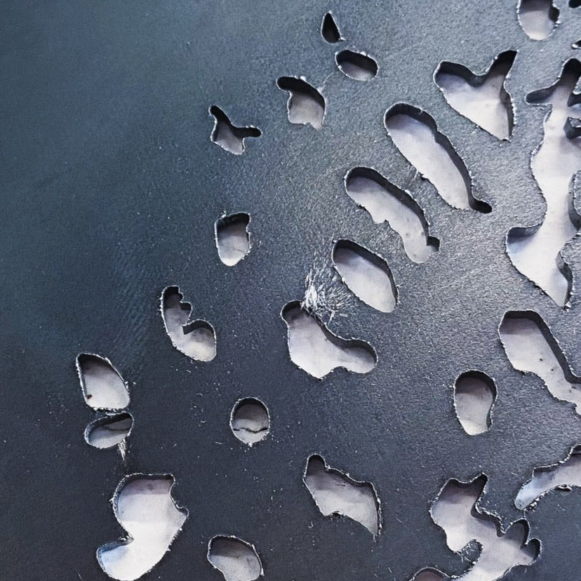 Close-up of Metalbird Seconds by Metalbird Canada - Seconds: a dark metal surface with organic-shaped cutouts revealing lighter material below. The smooth, rounded edges create a striking silhouette reminiscent of modern yard art.