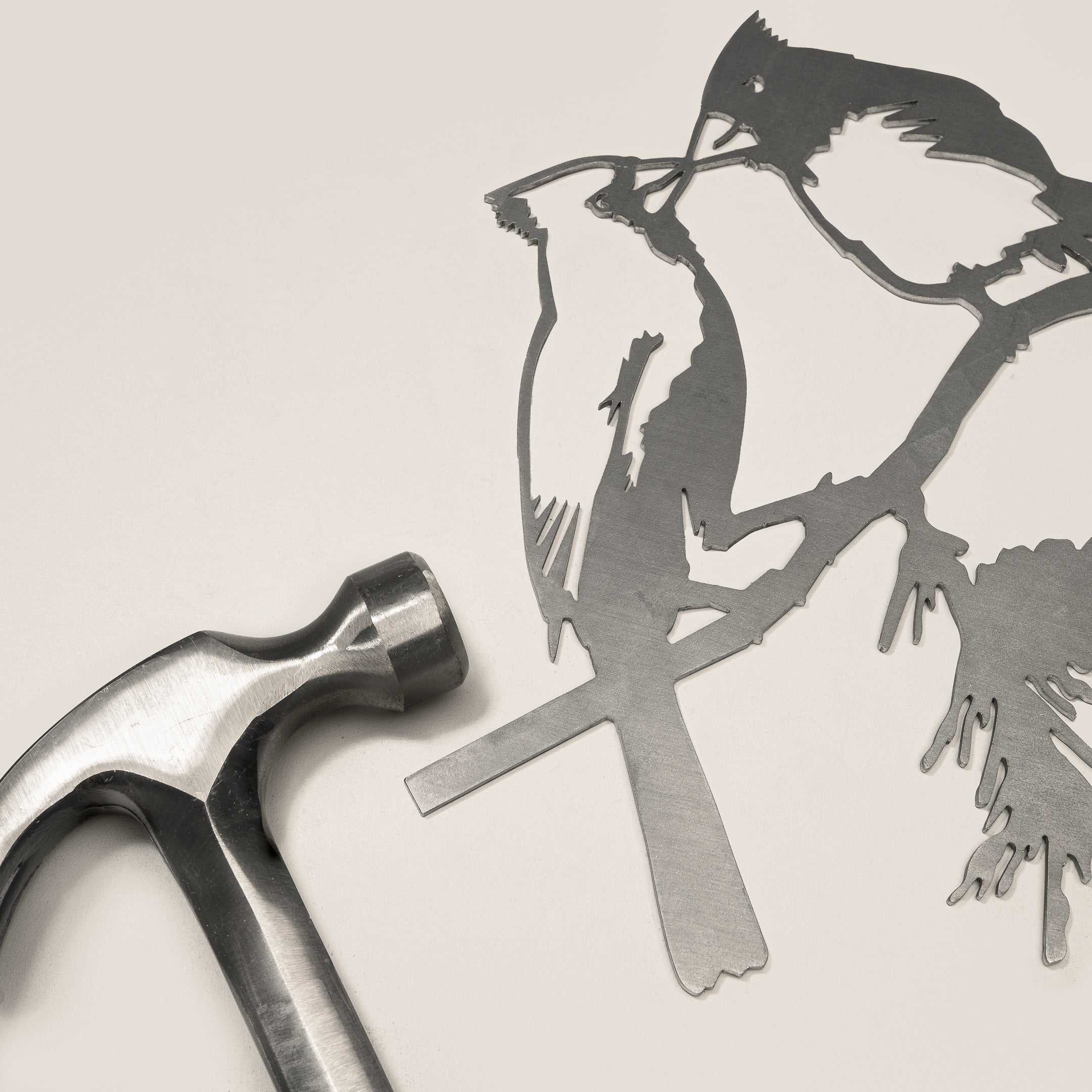 The Metalbird Canada Kissing Cardinals—a delicate metal silhouette of two birds on a branch—rests beside a metal hammer against a neutral background, offering perfect inspiration for garden art enthusiasts.