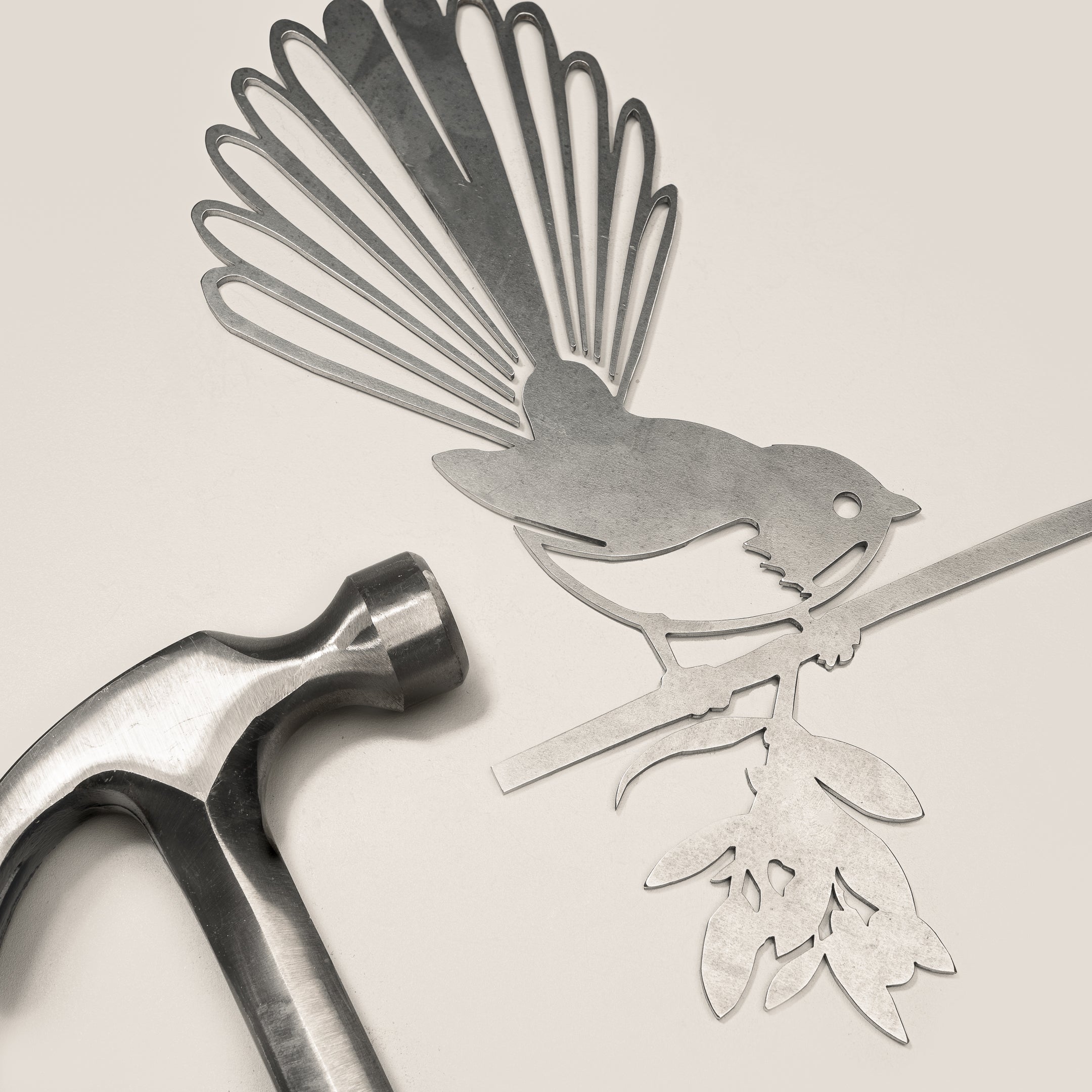 The Fantail / New Zealand Pīwakawaka by Metalbird Canada, a corten steel bird silhouette with floral branch, rests on a light surface beside a silver hammer—an ideal gift for nature lovers.
