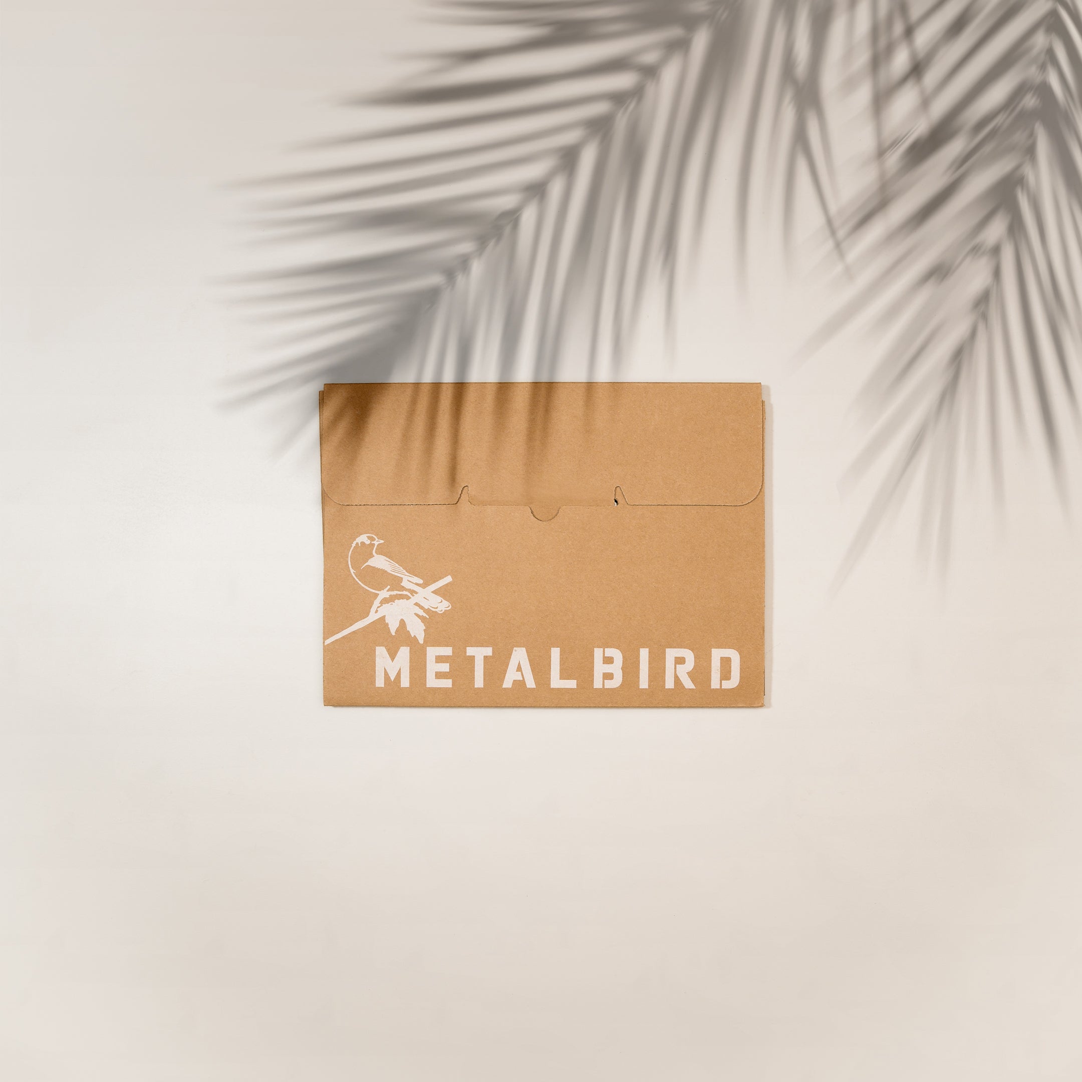 A brown envelope with a white illustration of a hummingbird on a branch and "Metalbird Canada" rests on a light surface, hinting at the unique corten steel Hummingbird inside, as palm leaf shadows cross the top right corner.