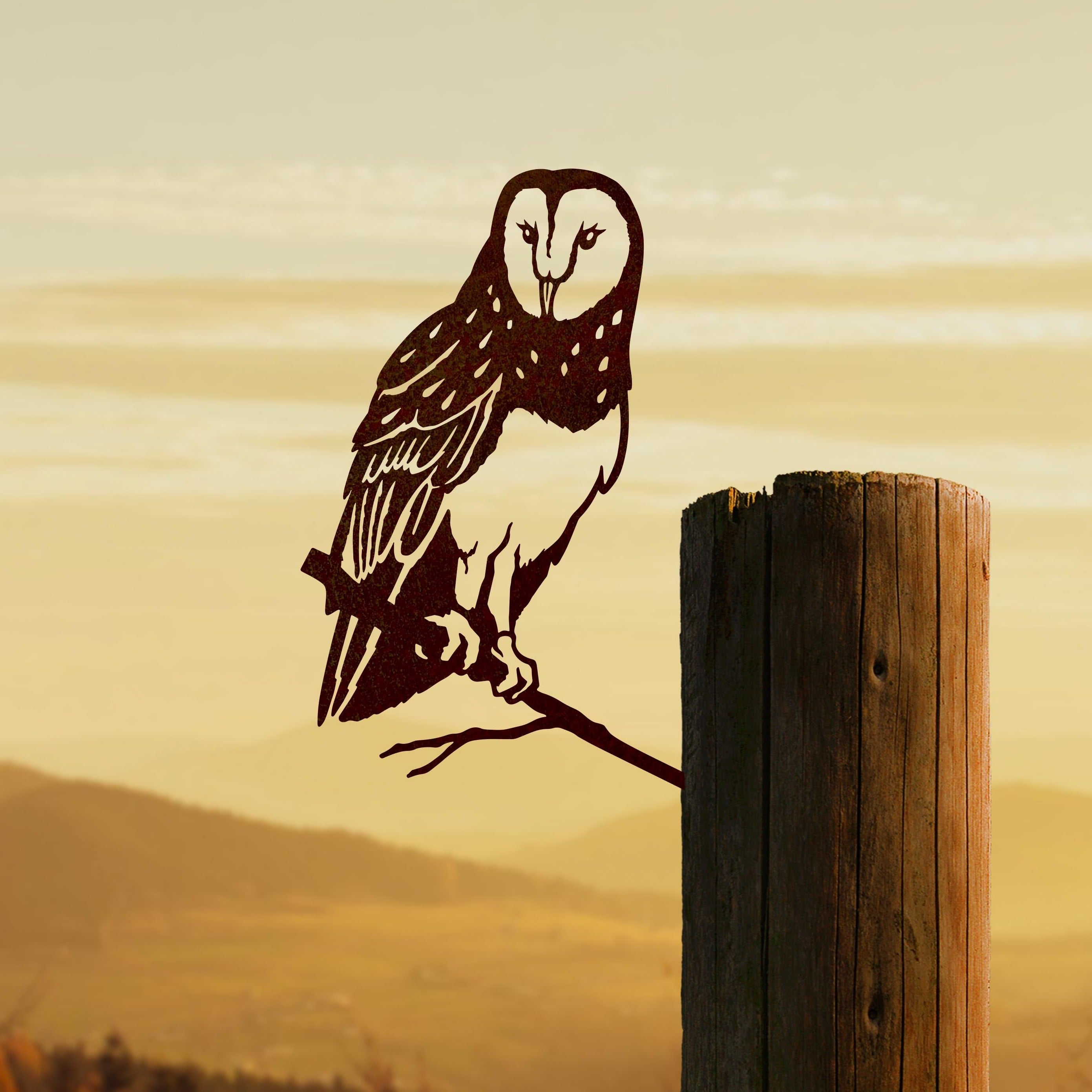 The Barn Owl by Metalbird Canada is a corten steel silhouette of an owl on a branch, mounted on a wooden post, pictured with hills and a golden sunrise or sunset sky in the background.