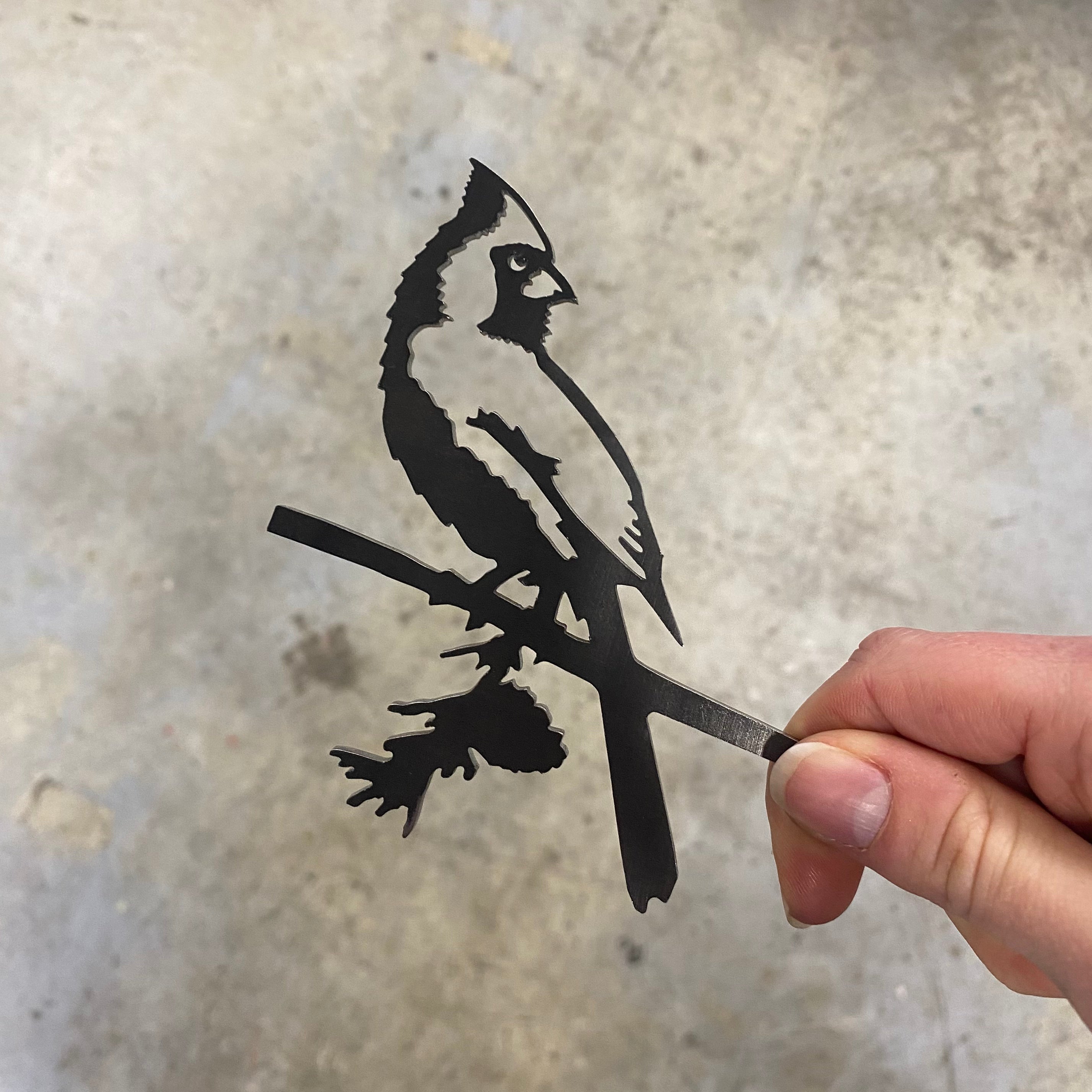 A hand holds the Mini Cardinal by Metalbird Canada, a cutout silhouette inspired by garden art in metal or corten steel, shown against a blurred gray background.