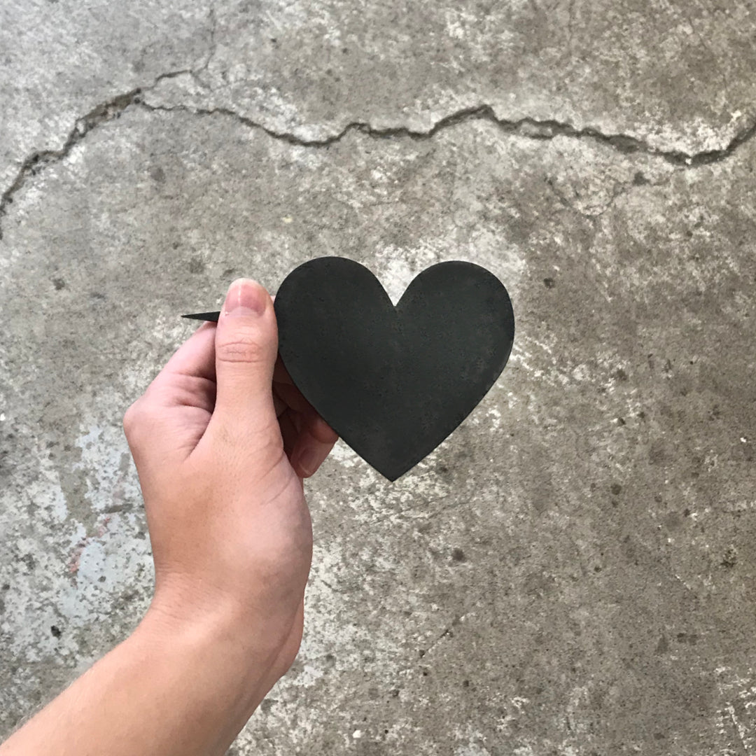 A hand holds the Metalbird Canada Mini Heart—a flat, black, heart-shaped silhouette—against rough concrete, making it perfect as unique garden or yard art.