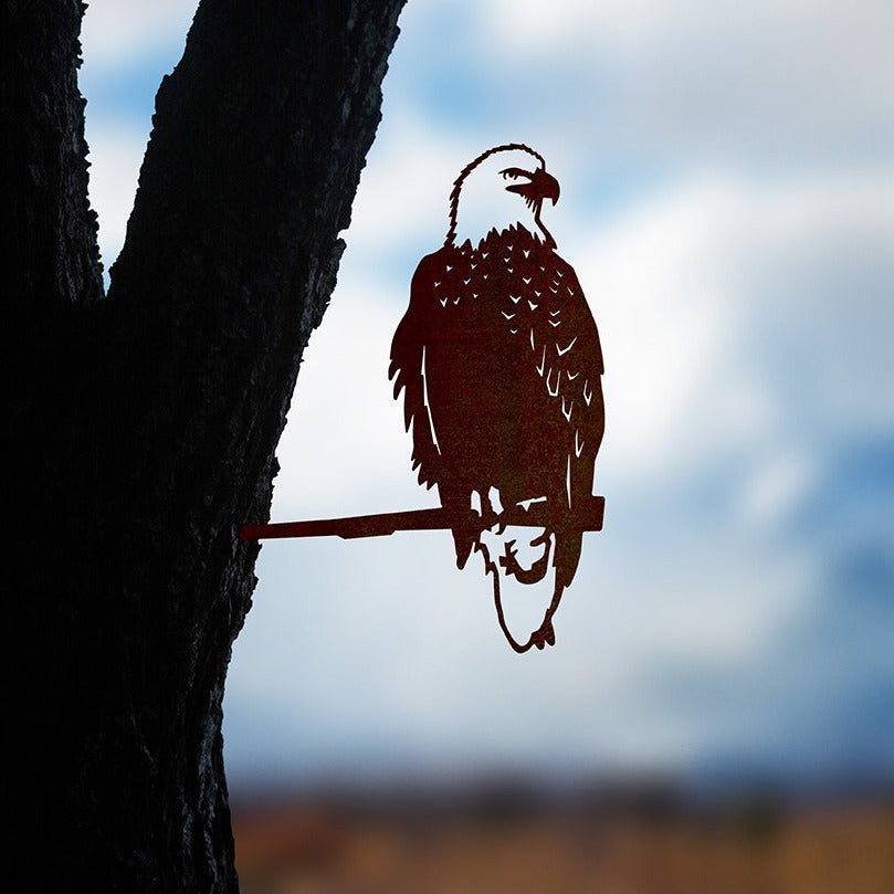 The Bald Eagle by Metalbird Canada is a corten steel silhouette of an eagle on a branch, mounted to a tree trunk to create eye-catching garden art with the sky and landscape softly blurred in the background.