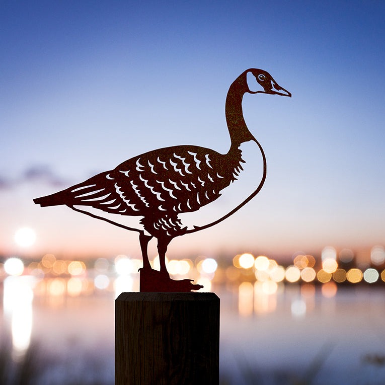 The Canada Goose by Metalbird Canada is a corten steel goose sculpture on a post, set against a blurred cityscape with colorful lights reflecting on water at dusk.