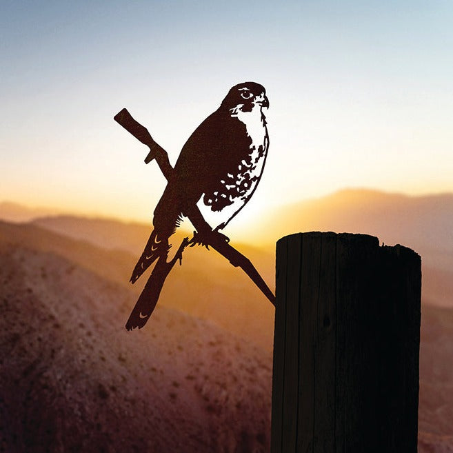 A Metalbird Canada Peregrine Falcon corten steel silhouette perches on a stick atop a wooden post, set against mountains at sunset with a softly glowing sky in the background.