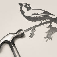The Cardinal Mom & Mini Set by Metalbird Canada—a metallic hammer and a metal cutout of a cardinal on a branch—rests on a light surface, making it an ideal gift for bird lovers.