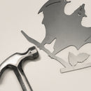 A Metalbird Canada Bat, a flat metal cutout with wings spread, perched on a branch with two leaves, lies beside a metal hammer—an ideal piece of yard art or a unique gift set against a light background.