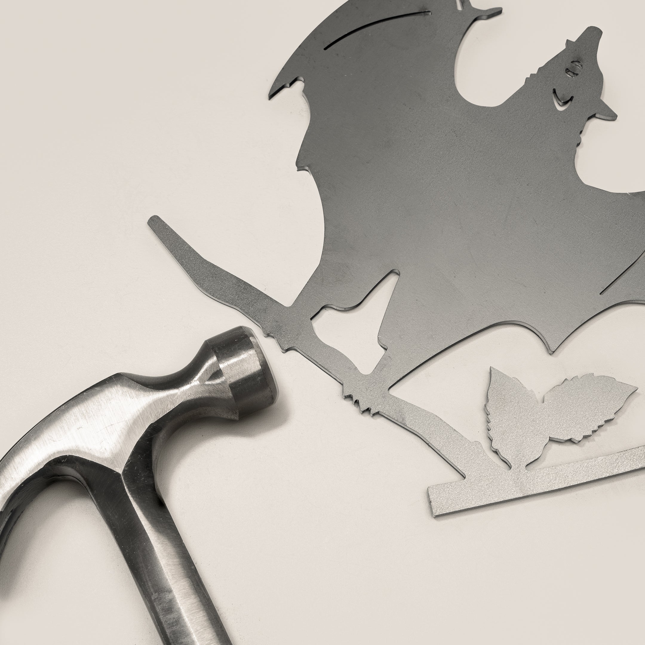 A Metalbird Canada Bat, a flat metal cutout with wings spread, perched on a branch with two leaves, lies beside a metal hammer—an ideal piece of yard art or a unique gift set against a light background.