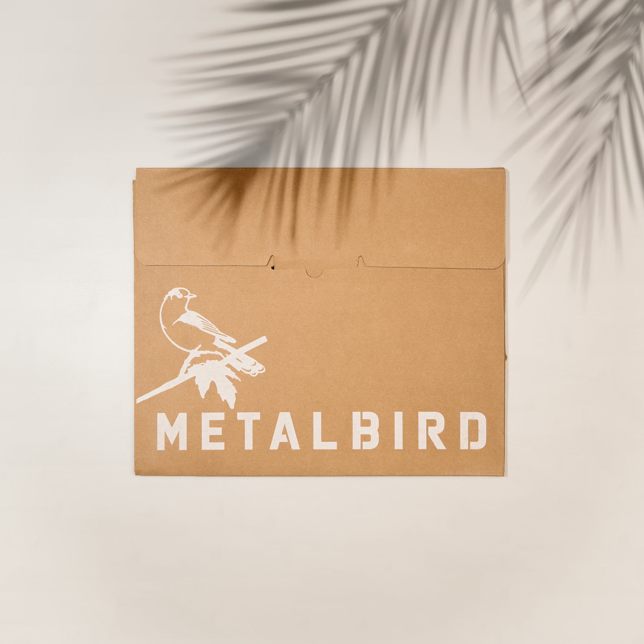 A brown cardboard envelope with a white silhouette illustration of a turkey and "Metalbird Canada" underneath, lying on a light surface with palm leaf shadows—perfect as a unique gift.