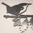 A MBPROMO Wren garden ornament made from Corten steel, featuring a bird perched on a leafy branch. Photographed on a light surface, the metal has a smooth, reflective finish and is shown from the side.