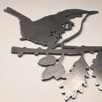 A MBPROMO Wren garden ornament made from Corten steel, featuring a bird perched on a leafy branch. Photographed on a light surface, the metal has a smooth, reflective finish and is shown from the side.