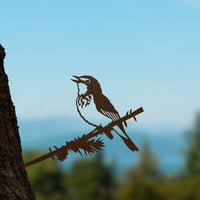 The Metalbird Canada Warbler silhouette by Metalbird Canada is mounted on a tree trunk with a blurred blue sky and green landscape behind it—a unique gift for bird conservation enthusiasts.