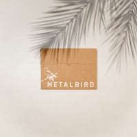 A brown cardboard envelope with "Metalbird Canada" and a bird graphic in white holds the Cardinal Mom & Mini Set—an ideal gift for bird lovers or moms—shown on a light surface with palm leaf shadows overhead.