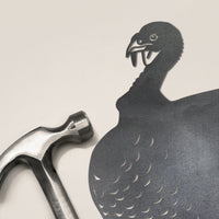 A metal hammer rests on a light background, casting a shadow that resembles the Metalbird Canada Turkey, with feathered detail and a hooked beak—evoking striking metal yard art.