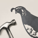 A metal hammer sits beside the Metalbird Canada Quail corten steel silhouette, creating unique yard art on a light-colored surface.