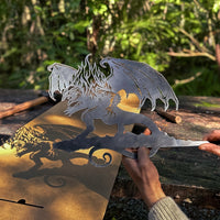 A person holds the BALROG™ metal dragon cutout by Metalbird Canada, its wings casting a striking shadow on cardboard outdoors amid lush greenery—an eye-catching piece of garden art.