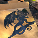 The BALROG™ by Metalbird Canada is a black metal dragon silhouette with wings and curled tail, displayed as striking garden art on cardboard backing in sunlight; mentions Middle-earth Enterprises and New Line.