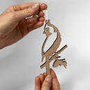 A person holds a Metalbird Canada Wooden Hanging Decoration shaped like a bird on a branch, ideal as garden art or a unique gift, suspended by string against a plain white background.