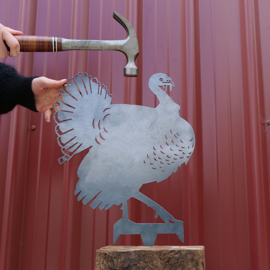 Someone holds the Metalbird Canada Turkey silhouette upright while another prepares to hammer it into place, set against a red metal wall that highlights this distinctive garden art piece.