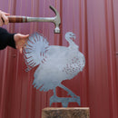 Someone holds the Metalbird Canada Turkey silhouette upright while another prepares to hammer it into place, set against a red metal wall that highlights this distinctive garden art piece.
