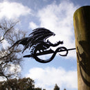 The FELL BEAST™ by Metalbird Canada—a black corten steel dragon silhouette—stands on a wooden post with a partly cloudy sky and tree branches in the background.