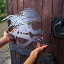 A person holds Metalbird Canada's FELL BEAST™ metal silhouette, featuring a dragon perched on a stylized base—unique garden art—displayed in front of a wooden door with iron hardware.