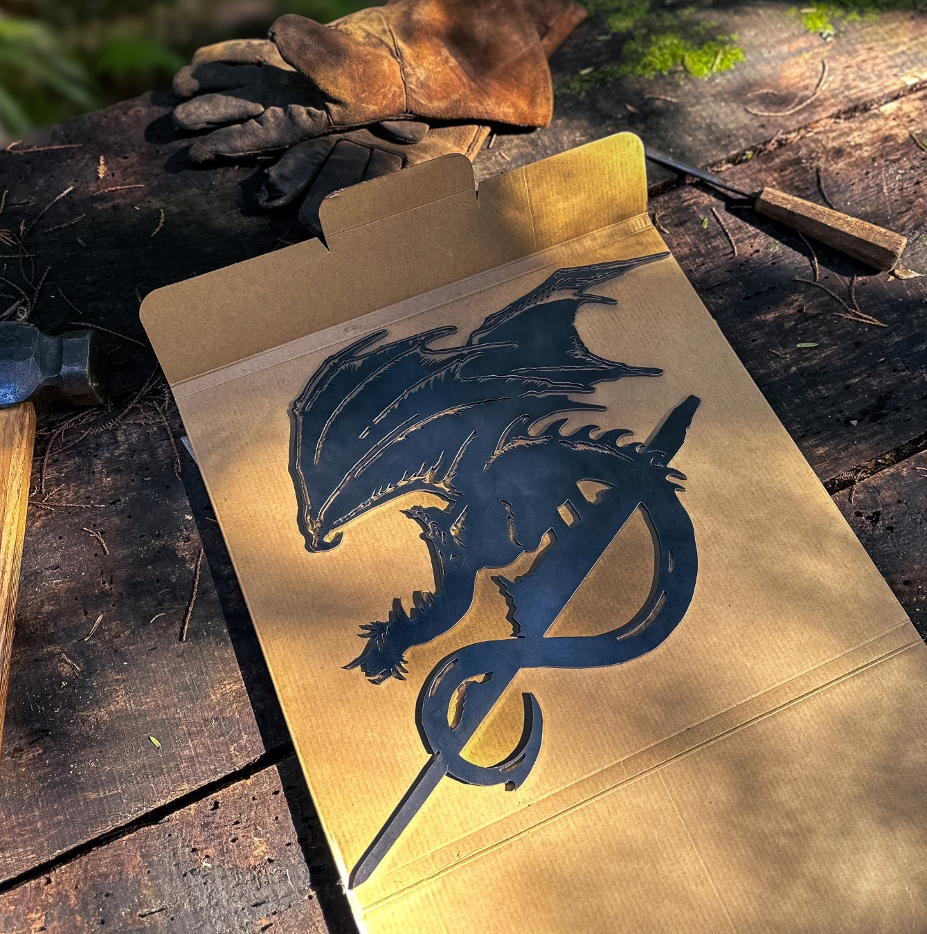 The FELL BEAST™ by Metalbird Canada is a black metal dragon silhouette with wings and a curled tail, resting on an open box beside tools—perfect as eye-catching garden or yard art for your outdoor space.