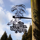 A corten steel THE GREEN DRAGON™ sign by Metalbird Canada, shaped like a dragon and featuring its name, hangs from a wooden post with trees and a blue sky with clouds in the background.