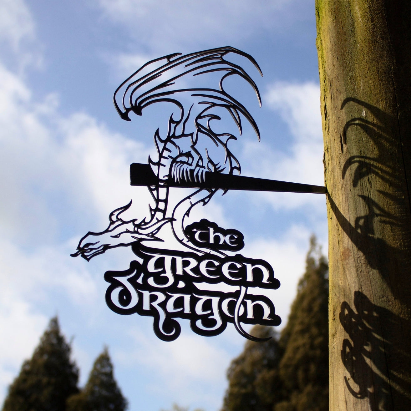A corten steel THE GREEN DRAGON™ sign by Metalbird Canada, shaped like a dragon and featuring its name, hangs from a wooden post with trees and a blue sky with clouds in the background.
