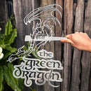 A hand holds the THE GREEN DRAGON™ metal silhouette sign by Metalbird Canada, featuring stylized lettering and displayed as unique garden art in front of a wooden fence and lush plants.