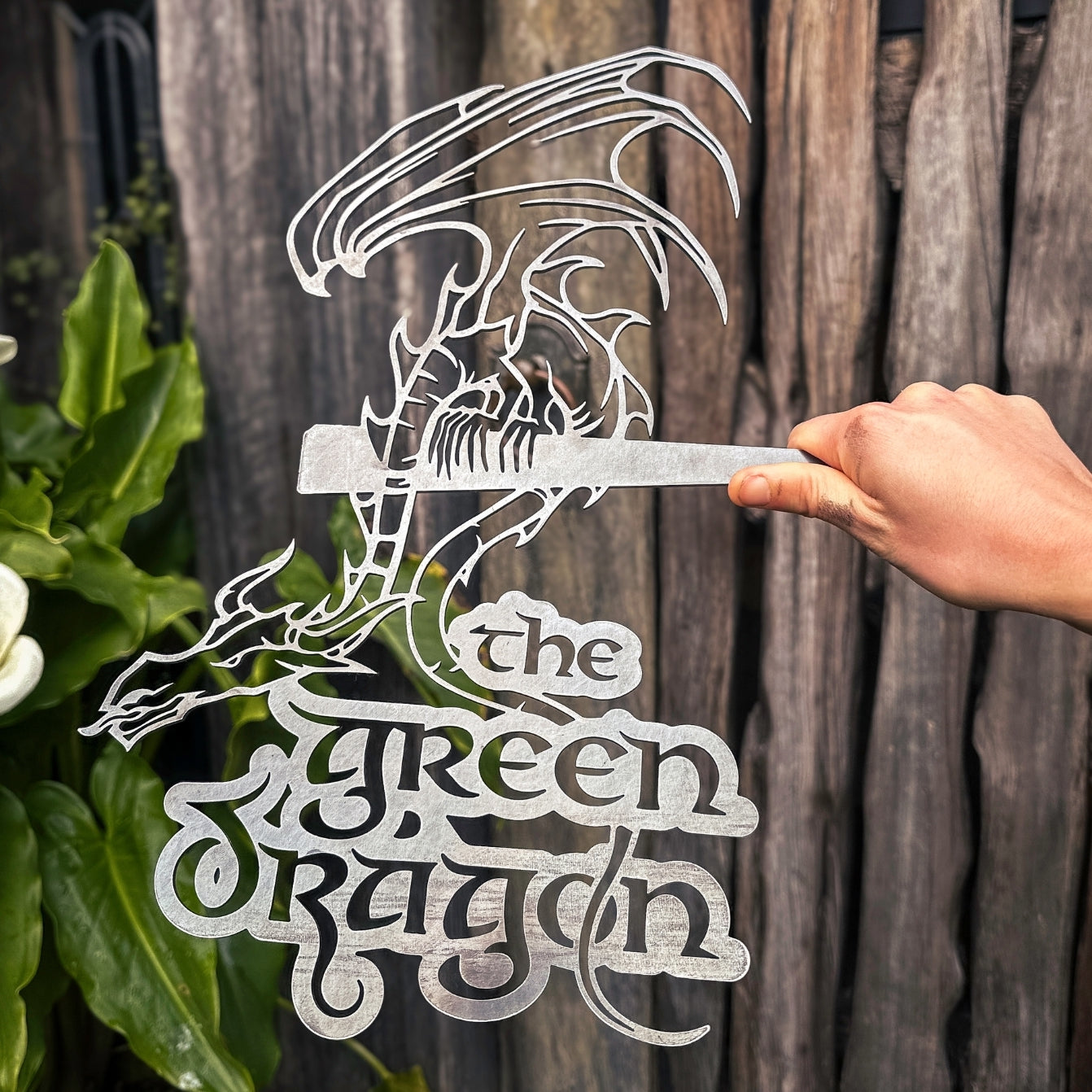 A hand holds the THE GREEN DRAGON™ metal silhouette sign by Metalbird Canada, featuring stylized lettering and displayed as unique garden art in front of a wooden fence and lush plants.