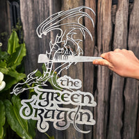 A hand holds the THE GREEN DRAGON™ metal silhouette sign by Metalbird Canada, featuring stylized lettering and displayed as unique garden art in front of a wooden fence and lush plants.
