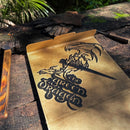 A cardboard box on rustic wood shows THE GREEN DRAGON™ by Metalbird Canada, featuring a dragon wrapped around a sword. Nearby gloves rest on planks, sparking inspiration for corten steel yard art in sunlit greenery.