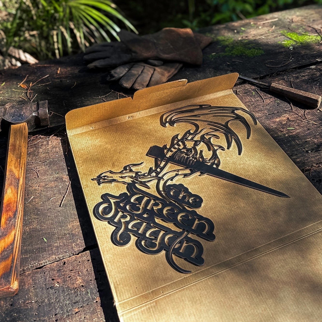 A cardboard box on rustic wood shows THE GREEN DRAGON™ by Metalbird Canada, featuring a dragon wrapped around a sword. Nearby gloves rest on planks, sparking inspiration for corten steel yard art in sunlit greenery.