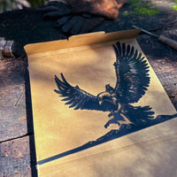 A cardboard box from Metalbird Canada, labeled "GANDALF™ ON GWAIHIR™," lies open on a weathered wooden surface with sunlight and shadows playing over it. The black eagle-themed illustration recalls motifs seen in corten steel garden art.