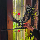 A hand holds Metalbird Canada's GANDALF™ ON GWAIHIR™ corten steel silhouette in front of a stained glass window with red accents; sunlight and green plants shine through, creating a unique gift.