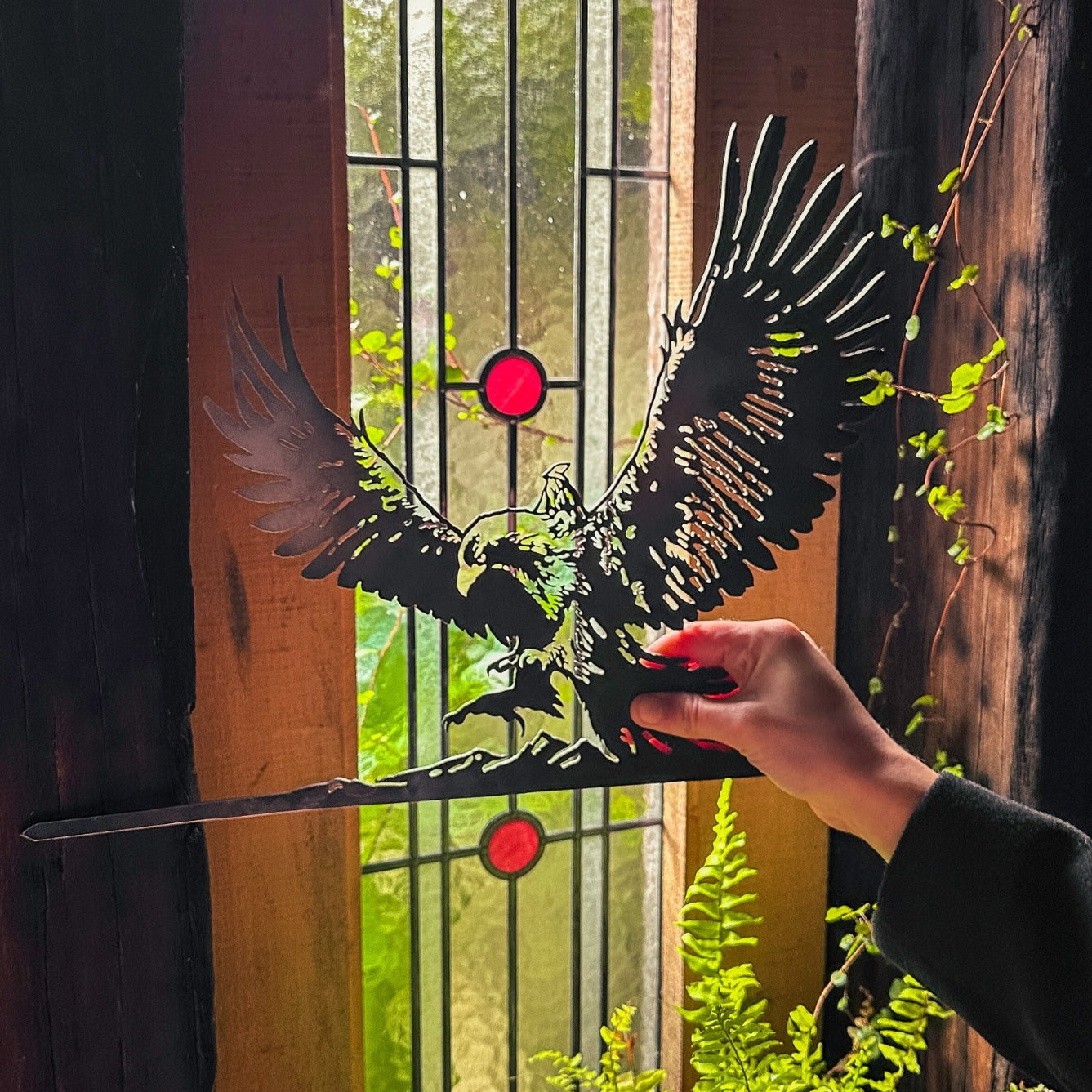 A hand holds Metalbird Canada's GANDALF™ ON GWAIHIR™ corten steel silhouette in front of a stained glass window with red accents; sunlight and green plants shine through, creating a unique gift.